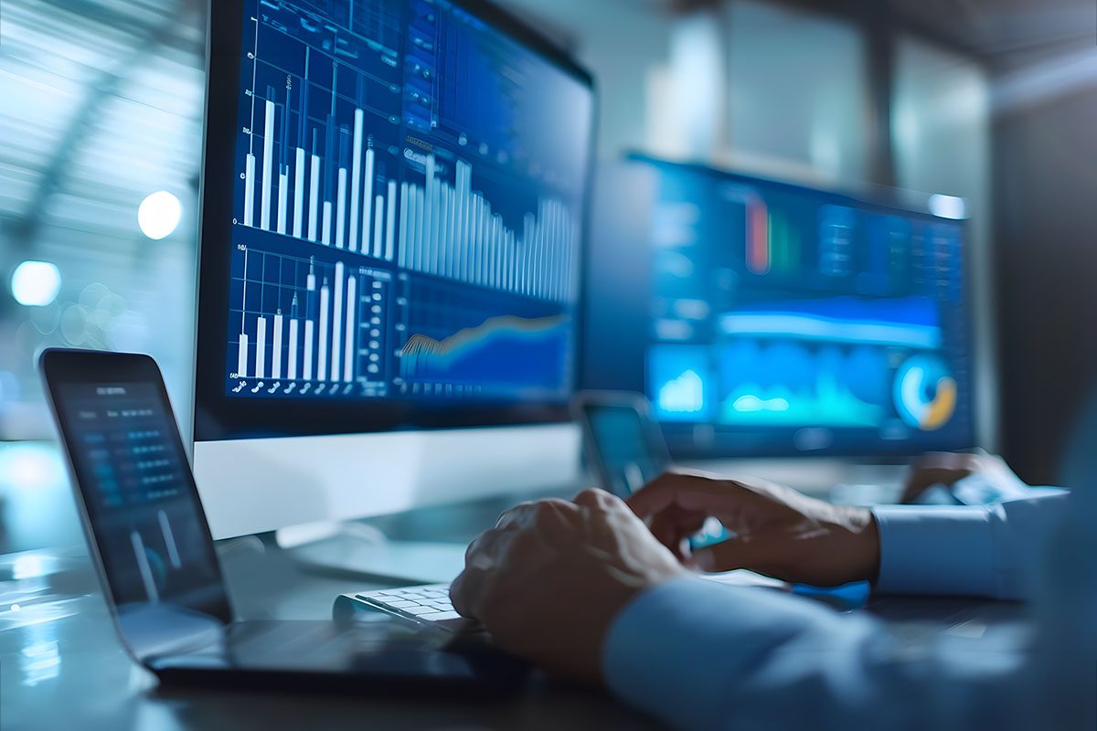 Person working at a desk with multiple monitors displaying data charts, graphs, and analytics in a modern office setting.