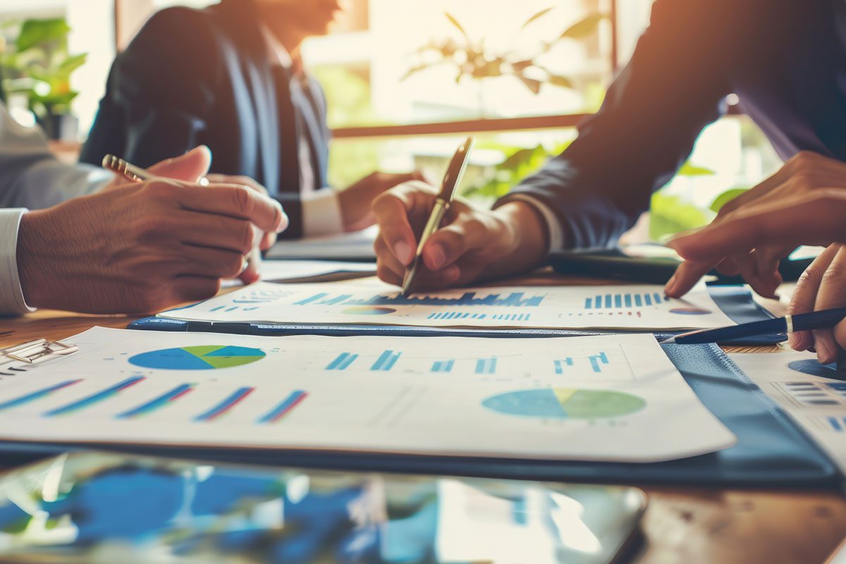 Three people review and discuss financial charts and graphs on paper at a table, with documents and pens visible in a well-lit office environment.