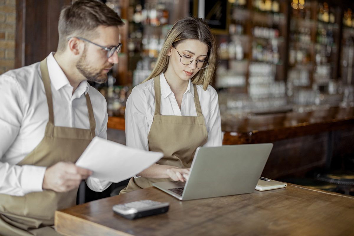 Two people wearing aprons sit at a bar counter; one looks at documents while the other types on a laptop. Shelves with bottles are visible in the background.