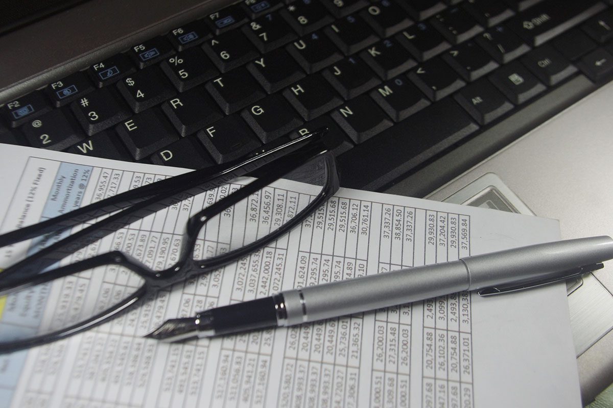 A pair of glasses and a silver pen rest on financial documents placed on a laptop keyboard.