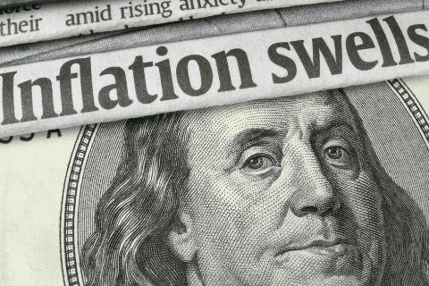Close-up of a newspaper headline reading Inflation swells placed over part of a U.S. hundred-dollar bill featuring Benjamin Franklins portrait.