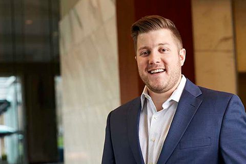 A man with short light brown hair and a beard, wearing a blue suit and white shirt, smiles while standing indoors in a modern office setting.