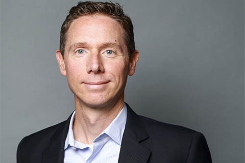 A man in a business suit with short brown hair stands in front of a plain gray background, looking at the camera with a neutral expression.