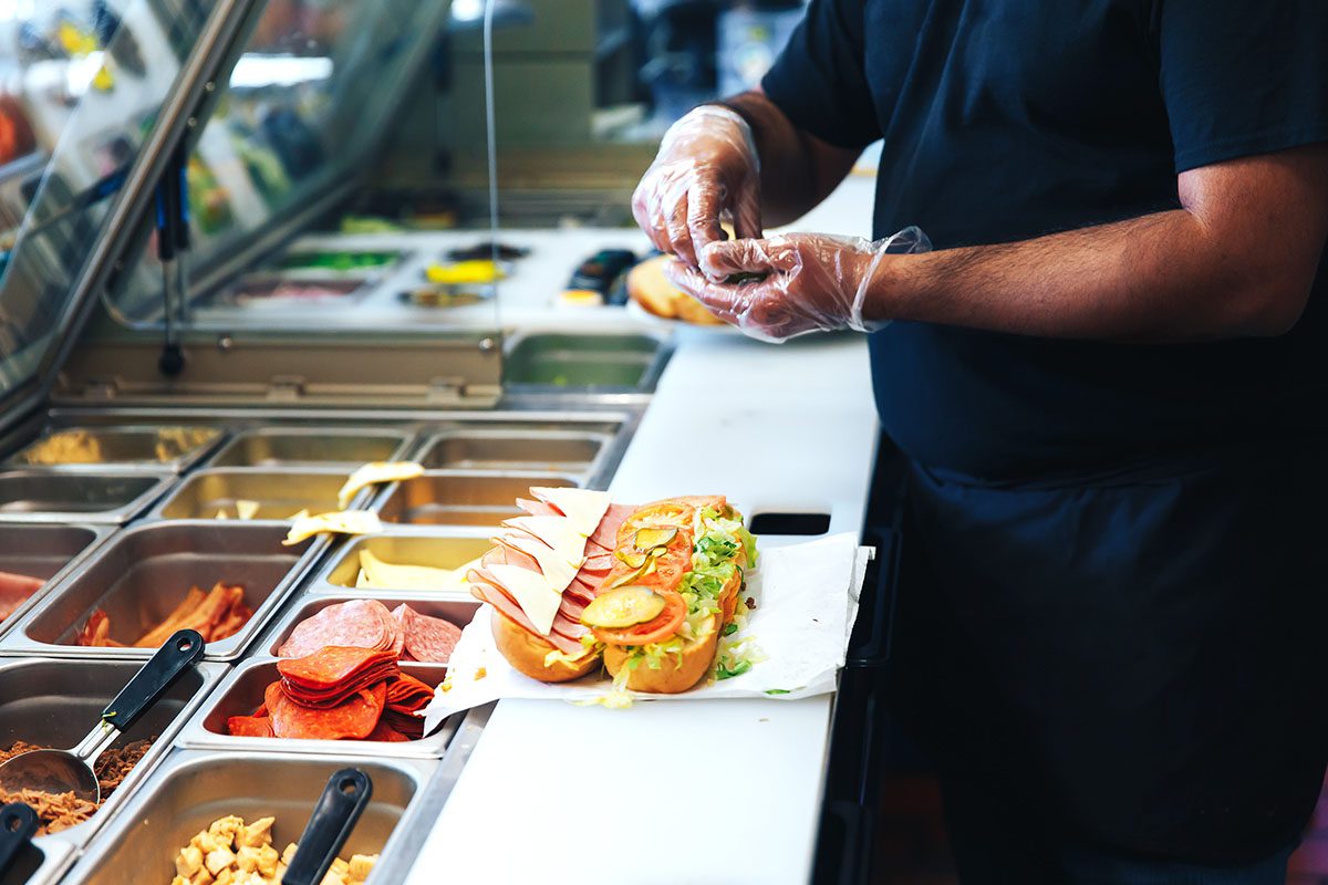 A person wearing gloves prepares a sub sandwich behind a counter with various deli ingredients in containers.