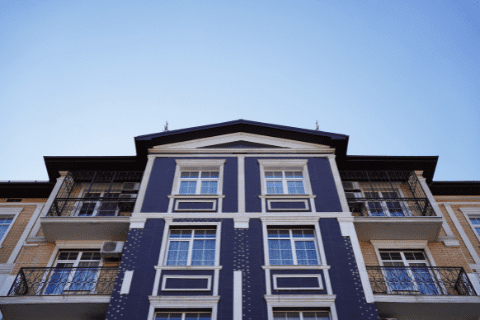 Facade of a modern apartment building with blue and white detailing, multiple balconies, and large windows, viewed from below against a clear blue sky.