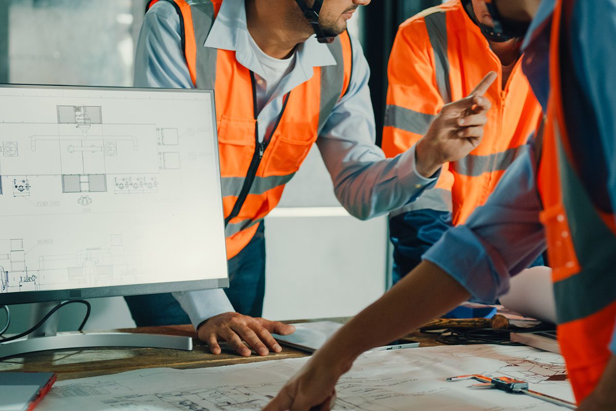 Three people in safety vests review architectural blueprints and plans on a table and a computer monitor in an office or construction site setting.