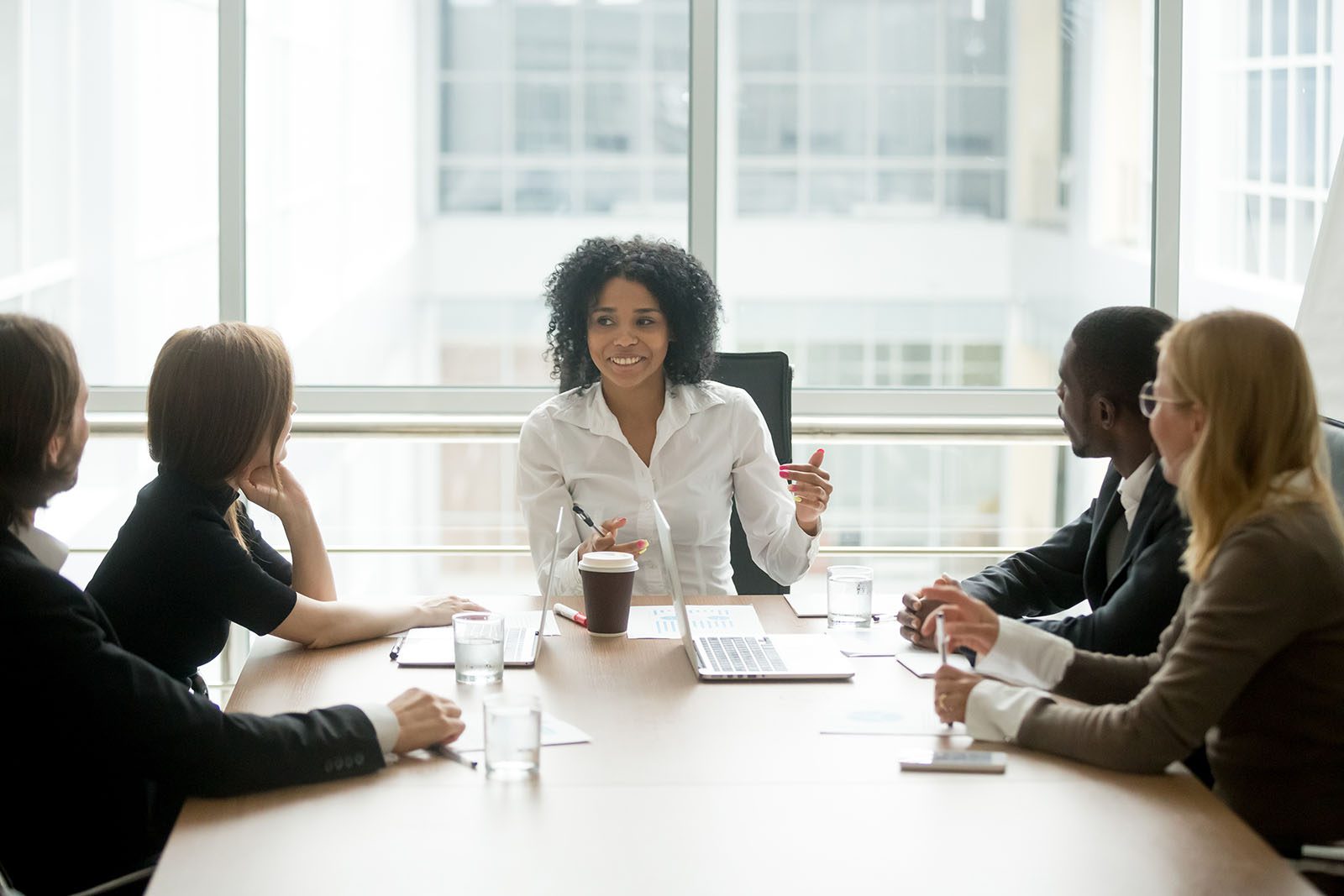 A woman leads a business meeting with four colleagues, seated around a conference table in a modern office with large windows.