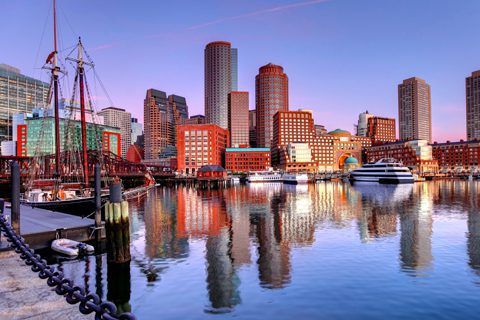 Boston Harbor waterfront with modern buildings, a historic sailing ship, and yachts reflected in the calm water at sunset.