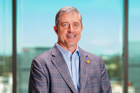 Man in a checked blazer smiles in front of a bright window with a blurred city view