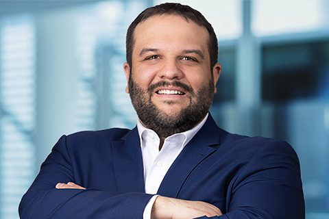 A man with short dark hair and a beard, wearing a navy suit and white shirt, stands smiling with arms crossed in front of a blurred office background.