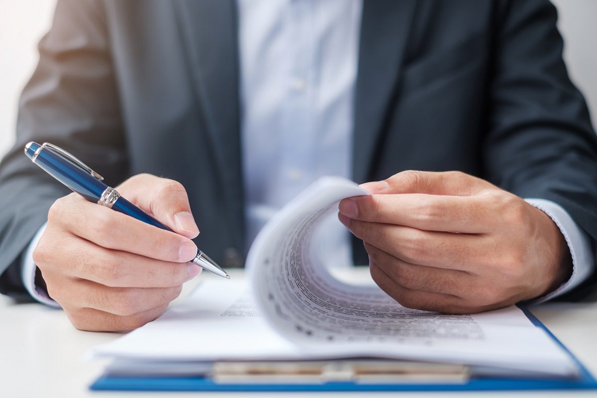A person in a suit holds a pen in one hand and flips through a stack of documents on a clipboard with the other hand.