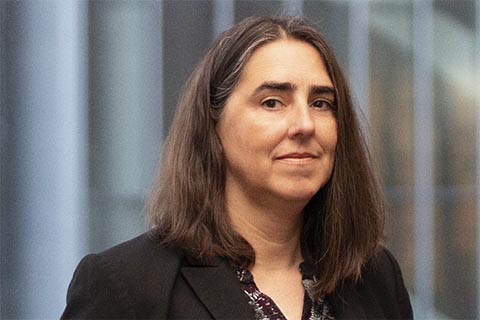 A woman with straight brown hair, wearing a dark blazer and patterned blouse, standing in front of a blurred background with vertical lines.