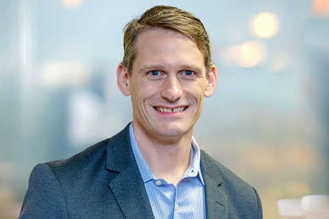 A man in a gray suit jacket and blue shirt smiles at the camera in an indoor setting with a blurred background.