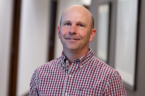 Man in a red checked shirt smiles in a blurred hallway