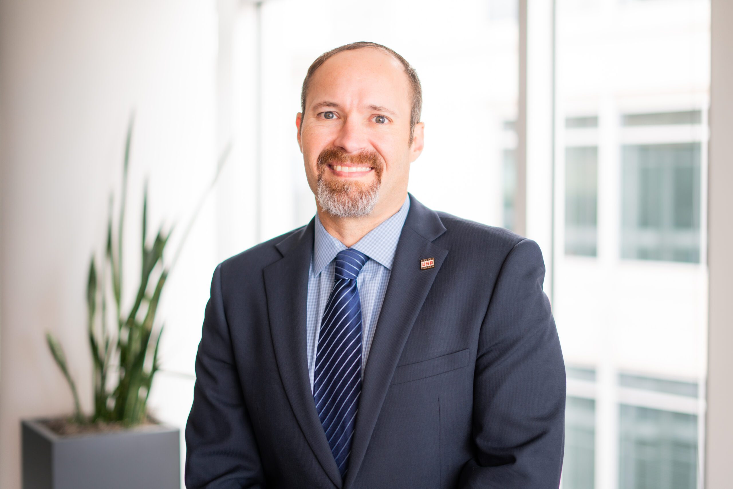 A man in a suit and tie stands indoors in front of large windows, with a potted plant visible in the background.