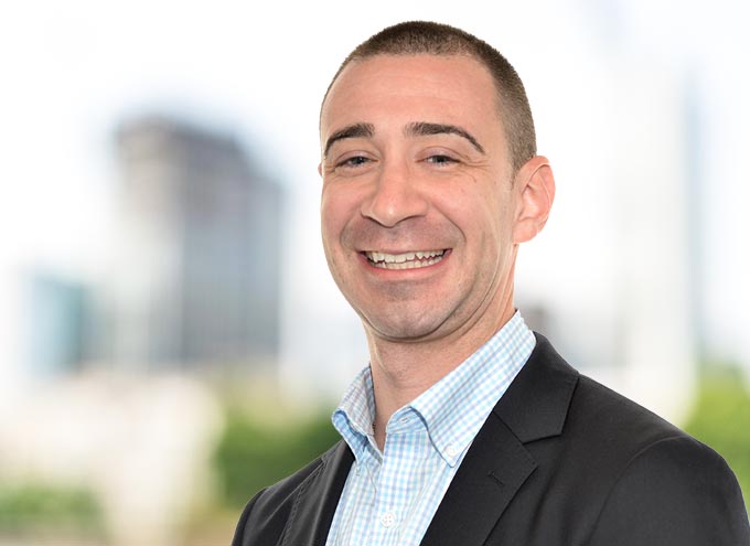 A man in a suit jacket and checked shirt smiles at the camera, with blurred buildings and greenery in the background.