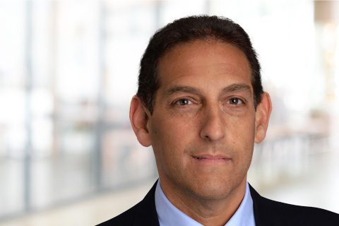 A man in a suit and tie stands in front of a blurred indoor background, facing the camera with a neutral expression.