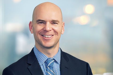 A bald man in a suit and blue checkered shirt smiles at the camera against a blurred indoor background.