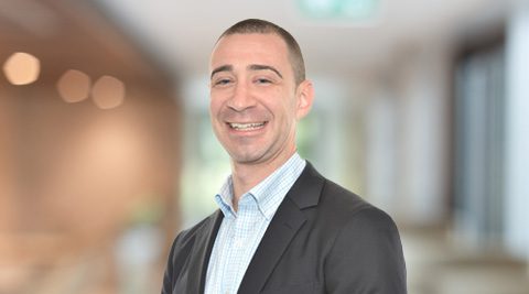 A man in a suit jacket and checkered shirt stands smiling in a blurred indoor setting with soft lighting.