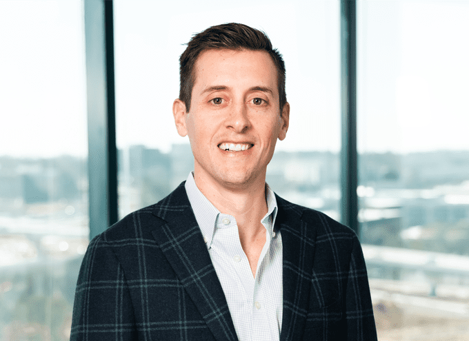 A man in a plaid blazer and light shirt stands indoors in front of large windows with an urban landscape in the background.