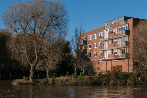 A red-brick apartment building with balconies stands by a riverbank, surrounded by leafless trees and greenery under a clear blue sky.