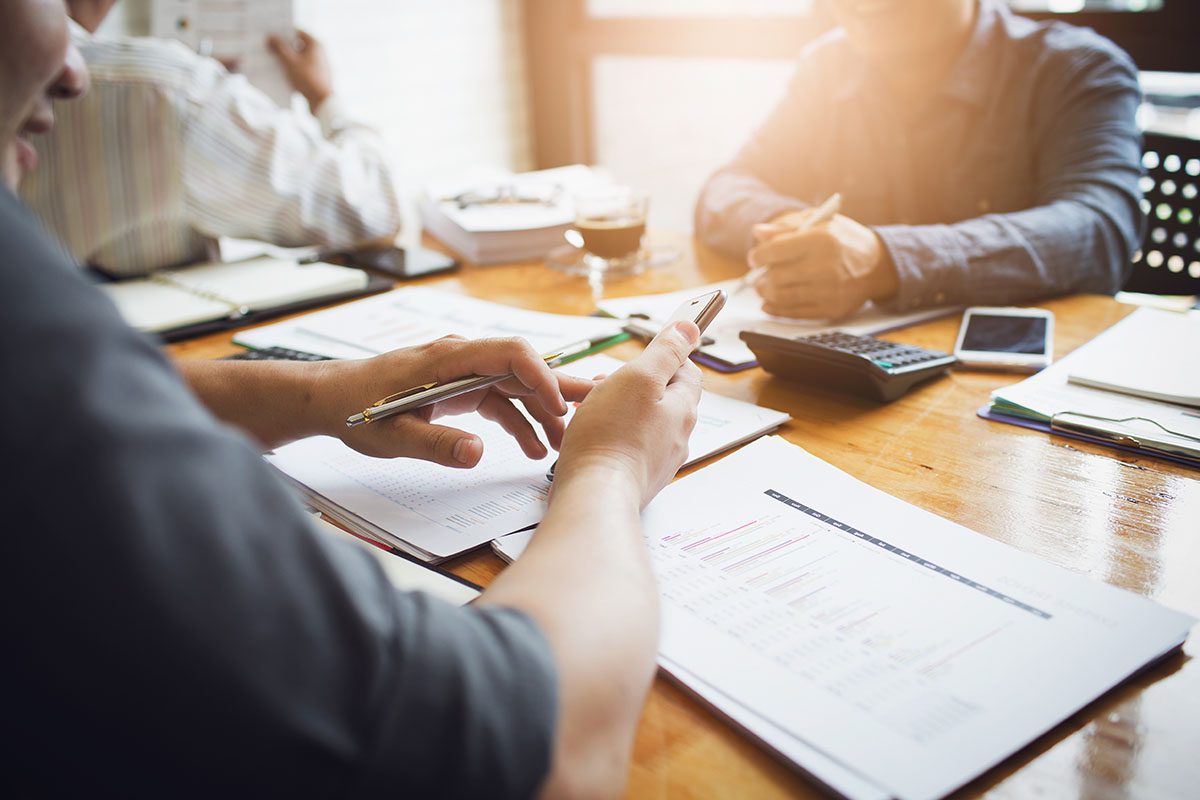 Three people sit at a table with documents, a calculator, notebooks, and a smartphone, discussing work in a bright office setting.