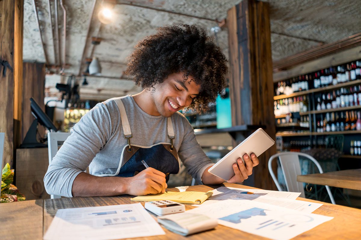 A person in an apron sits at a table in a restaurant, smiling while using a tablet and taking notes with papers, a calculator, and charts spread out in front of them.