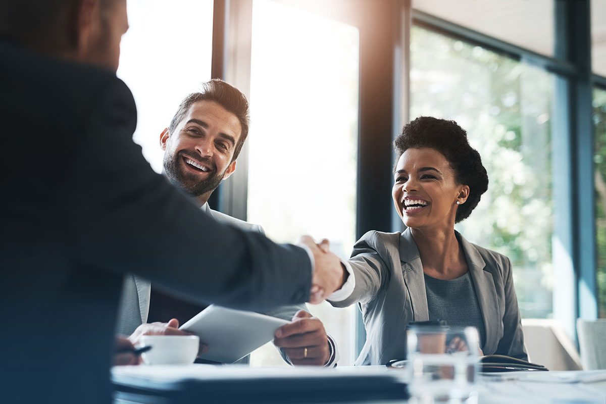Two people in business attire smile and shake hands across a table in a bright office setting, while a third person looks on.