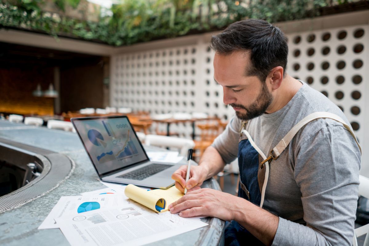 A man in an apron writes on a notepad at a counter with a laptop and printed charts in front of him in a modern indoor setting.