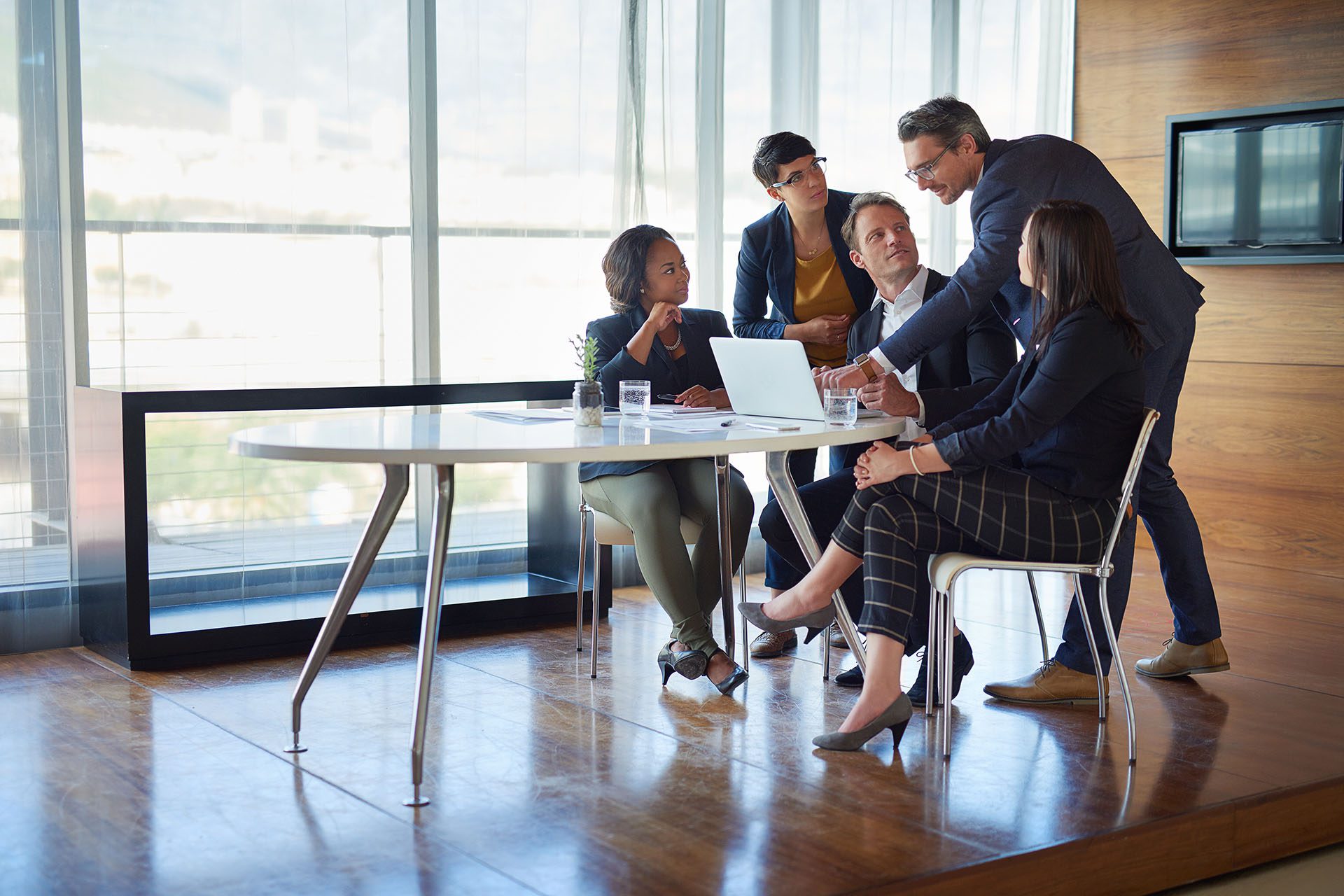 Five people in business attire sit and stand around a table in a modern office, discussing and looking at a laptop.