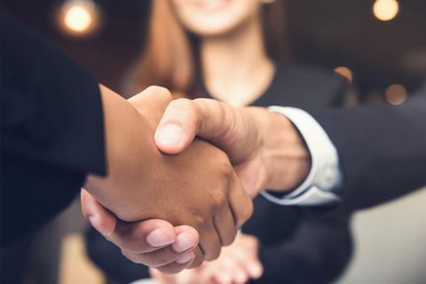Two people in business attire shaking hands, with a smiling person visible in the blurred background.