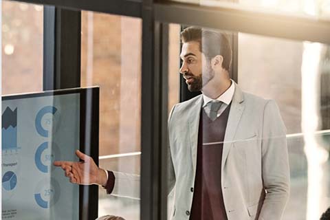 A man in a suit gestures toward a screen displaying charts and graphs during a presentation in an office setting.