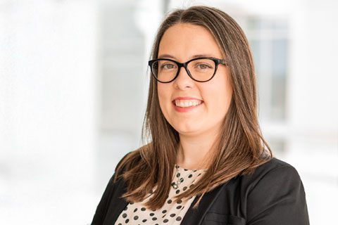 Woman with long brown hair and glasses, wearing a black blazer over a white blouse with black polka dots, smiling at the camera against a bright background.