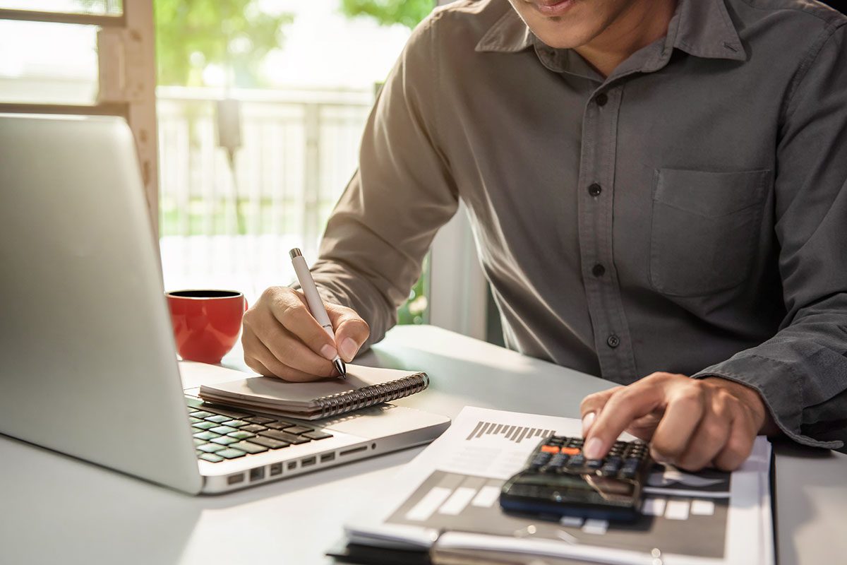 A person sits at a desk using a laptop, writing in a notebook, and pressing buttons on a calculator with papers and a red mug nearby.