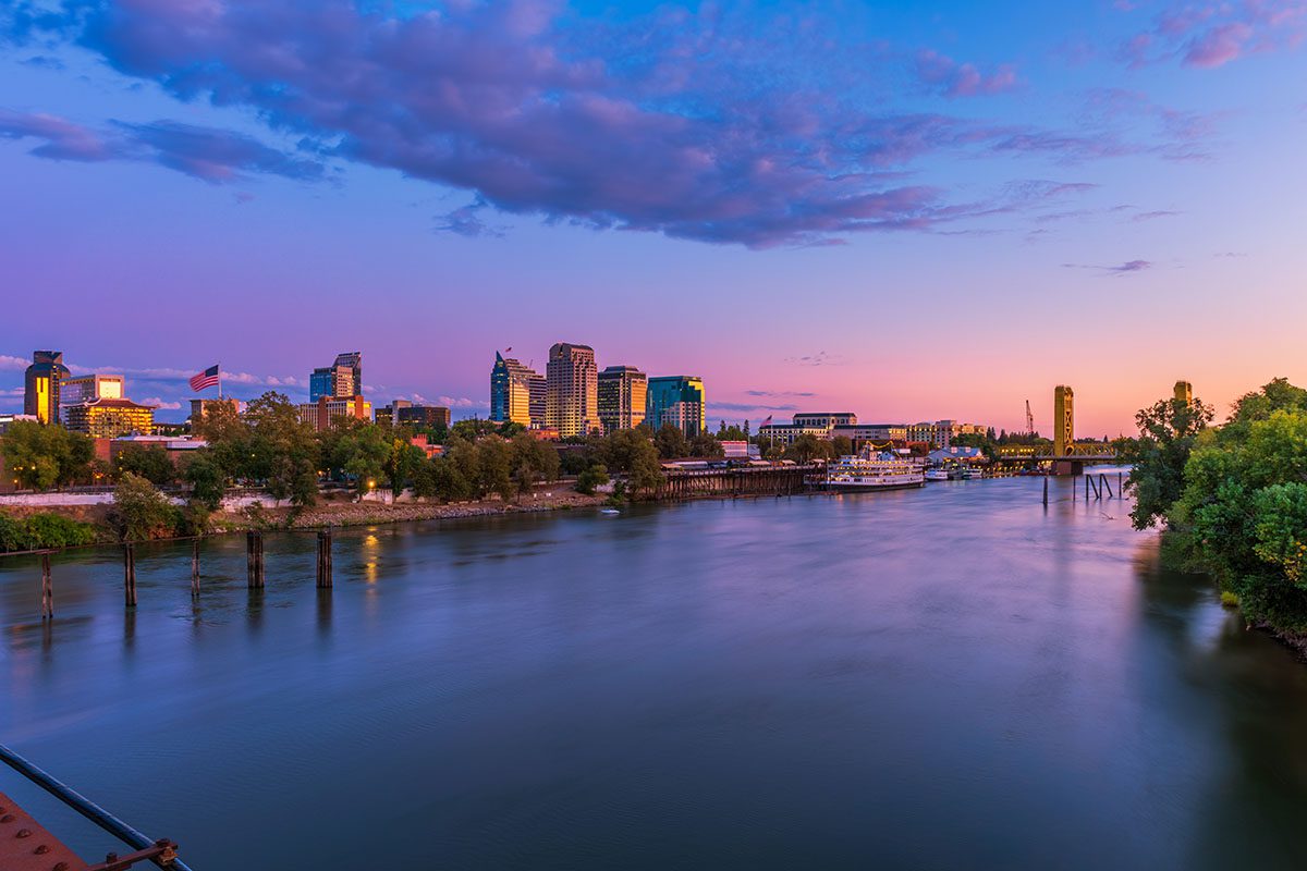 A wide river flows past a city skyline at sunset, with tall buildings, trees, and a tower visible under a colorful sky.