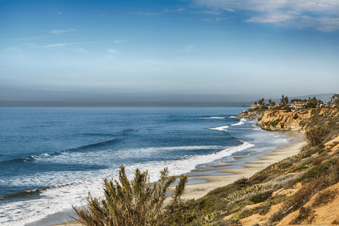 A sandy beach with gentle waves and cliffs under a clear blue sky, with houses visible on the distant shoreline.