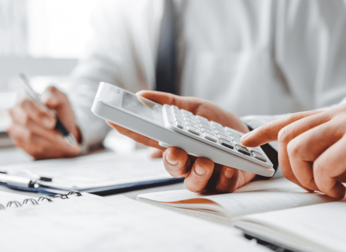 Two people working at a desk; one person uses a calculator while the other writes in a notebook. Papers and pens are visible.