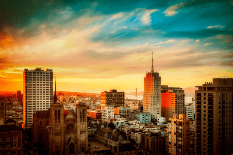 City skyline at sunset with tall buildings, a church in the foreground, and a bridge visible in the distance under a colorful sky.