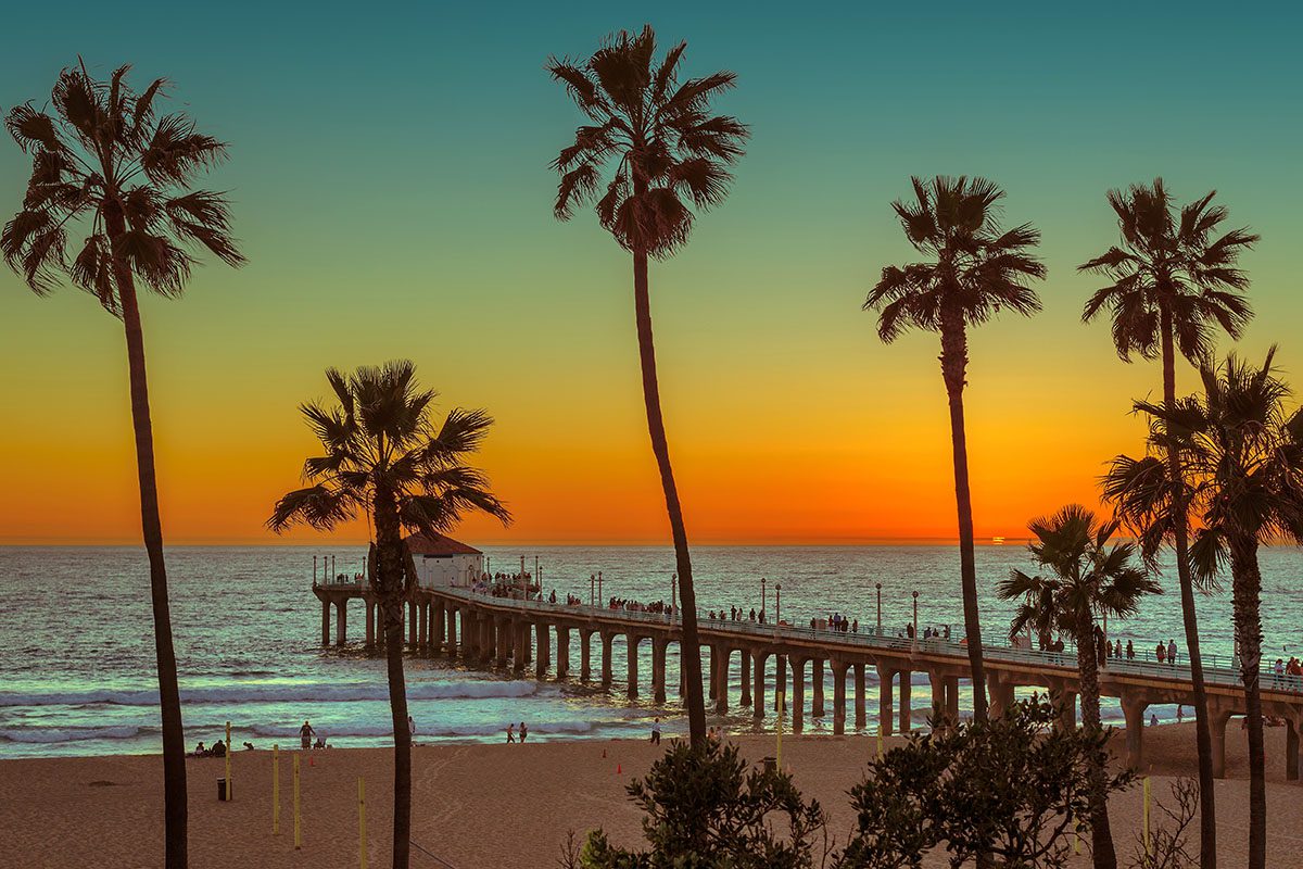 Palm trees frame a view of a pier extending into the ocean at sunset, with people walking along the pier and a colorful sky in the background.