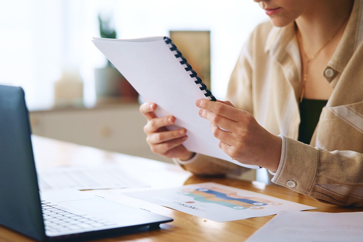 A person sits at a desk reading a spiral-bound report, with a laptop and printed charts in front of them.