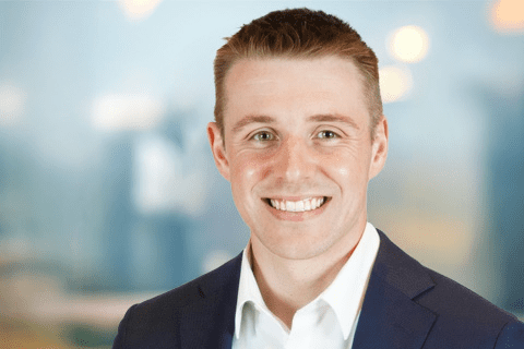 A man in a navy suit and white shirt smiles at the camera, standing in front of a blurred indoor background with soft lighting.