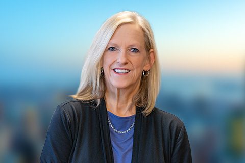 Blonde woman in a black cardigan smiles in front of a bright window with a blurred city view.