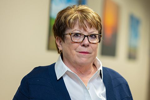 Short haired woman in glasses and a collared shirt smiles in a blurred hallway setting
