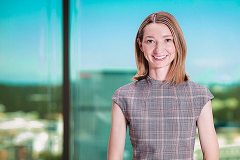 A woman with straight, shoulder-length hair wearing a plaid dress stands indoors, smiling, with a cityscape visible through large windows behind her.