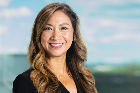 A woman with long wavy brown hair, wearing a black top, smiles at the camera with a blurred outdoor background.