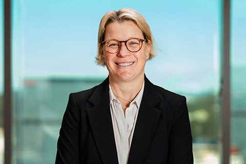 Blonde woman in glasses and a blazer smiles in front of a bright window with a blurred city view