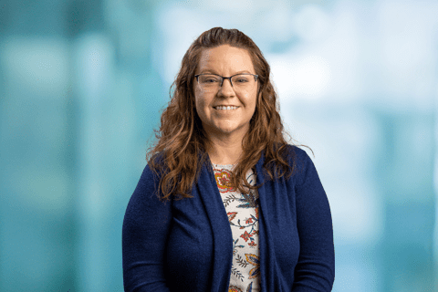 Woman with long brown hair and glasses, wearing a blue cardigan over a floral shirt, smiling at the camera against a blurred blue background.