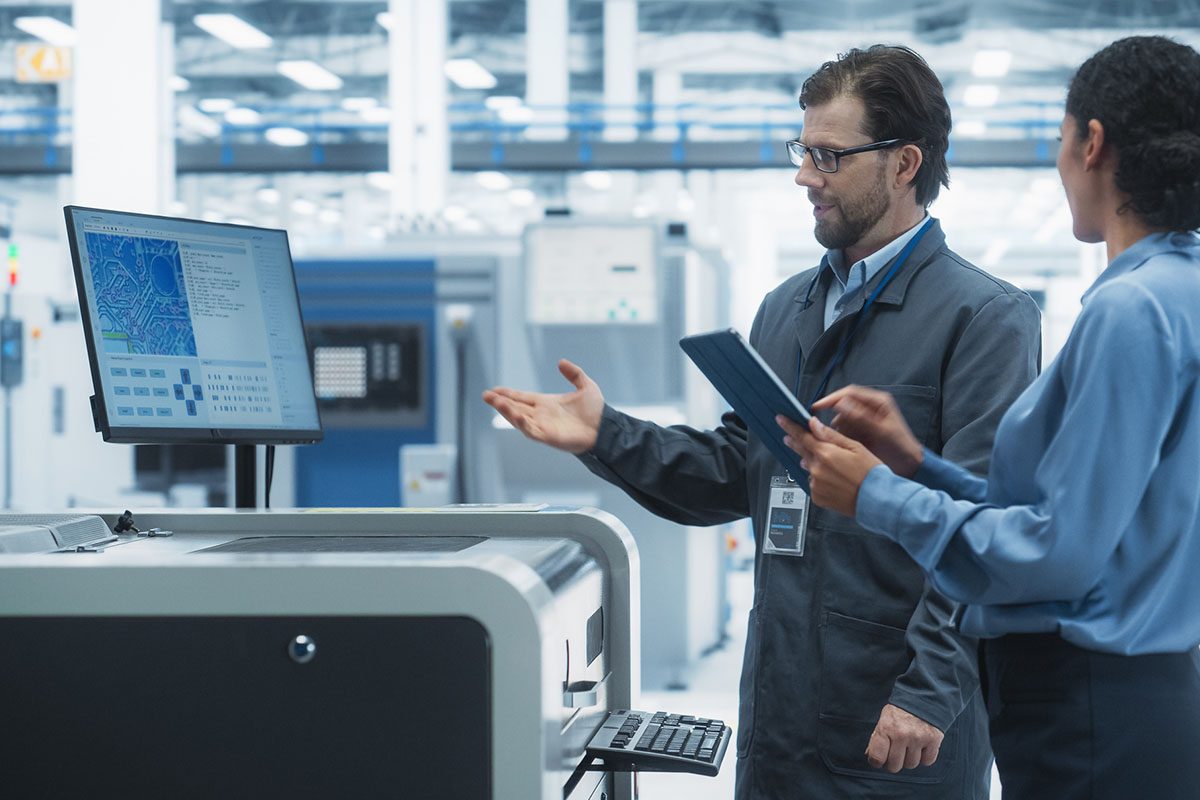 Two people in industrial attire discuss data displayed on a computer monitor in a high-tech manufacturing facility. One holds a tablet, the other gestures toward the screen.