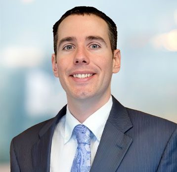 Man wearing a dark suit, white shirt, and patterned tie, smiling at the camera with a blurred light blue background.