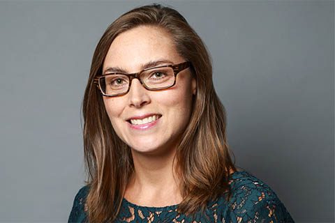 Woman with straight brown hair and glasses smiling, wearing a dark green lace top, posed in front of a plain gray background.